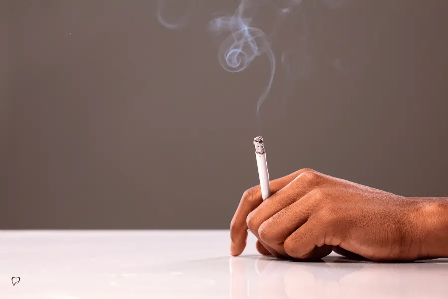 Main posée sur une table et tenant une cigarette allumée, d’où s’élève de la fumée.